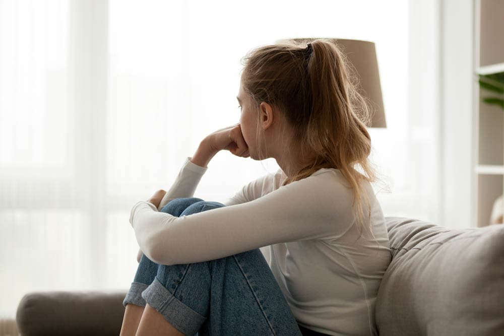 A young woman sitting on a couch looking thoughtful and distressed, symbolizing the emotional pain of rejection sensitivity often experienced with ADHD.