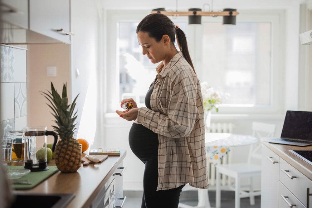 Pregnant woman in kitchen holding medication bottle and pills, reflecting decisions about ADHD medication use during pregnancy and child health outcomes.