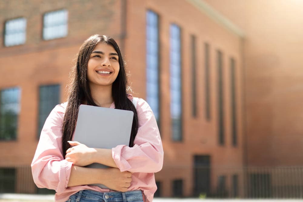 A smiling teenage student holding a laptop outside a school building, representing confidence and planning for post-high school options for teens with ADHD.