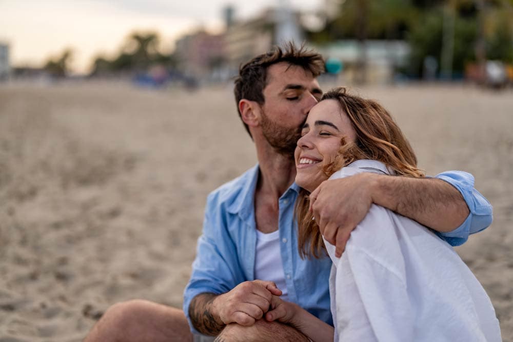 A man kissing a smiling woman on the forehead while holding hands on the beach, symbolizing the effects of ADHD on dating and romantic attraction.