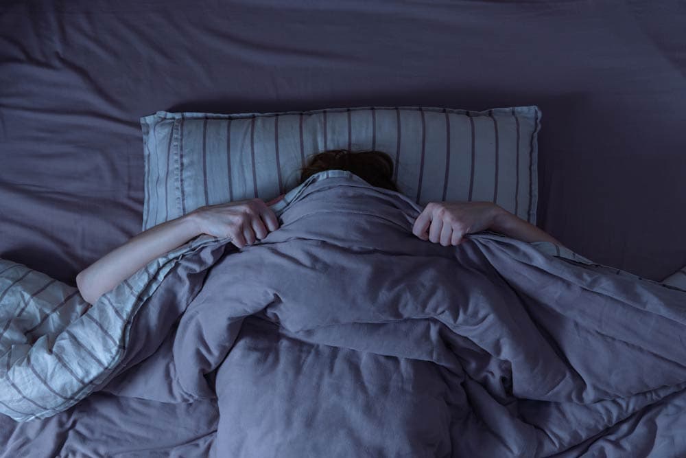 Person lying in bed under a blanket with only their hands visible, symbolizing how ADHD disrupts circadian rhythms and makes restful sleep difficult.