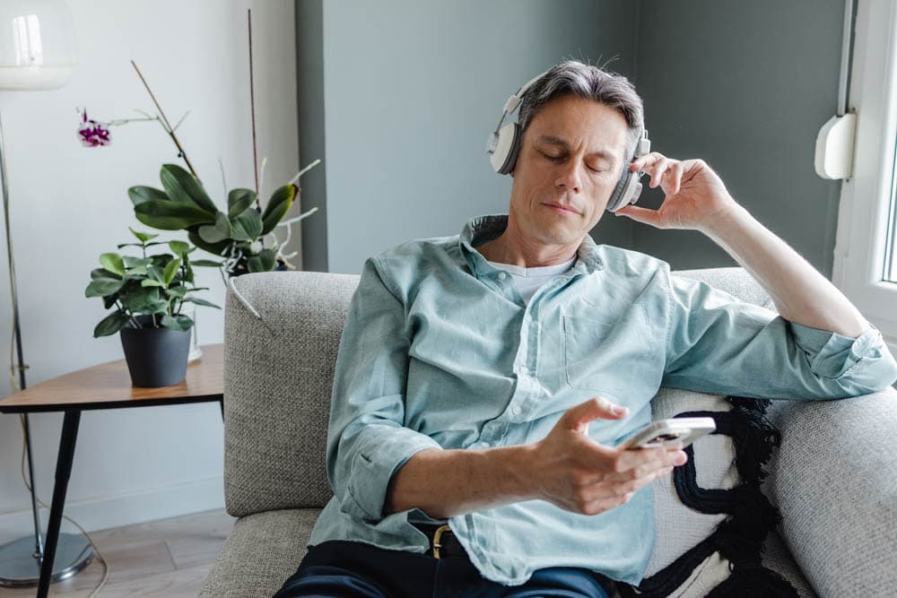 Middle-aged man sitting on a sofa with headphones, listening to a meditation app on his phone to practice mindfulness for ADHD focus and stress relief.
