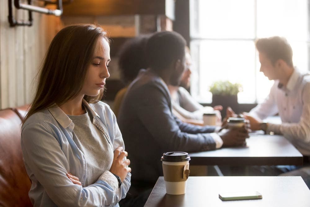 Young woman with ADHD sitting apart in a café, looking anxious while others socialize, representing the challenge of social anxiety and friendships.