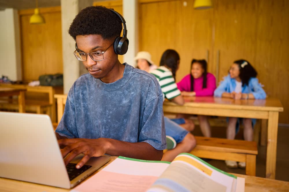 A college student with ADHD wearing headphones and focusing on a laptop while peers chat in the background, illustrating the importance of boundaries and saying no without guilt.