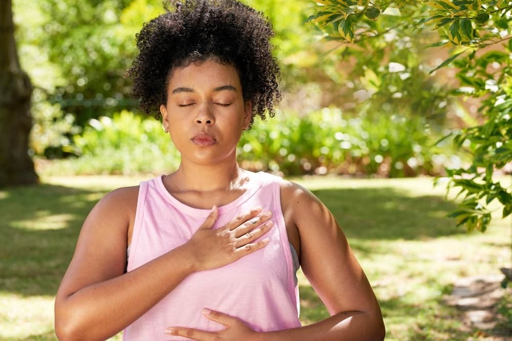 A woman practicing deep breathing outdoors with eyes closed and hands on her chest, representing stress relief and mindfulness for ADHD.