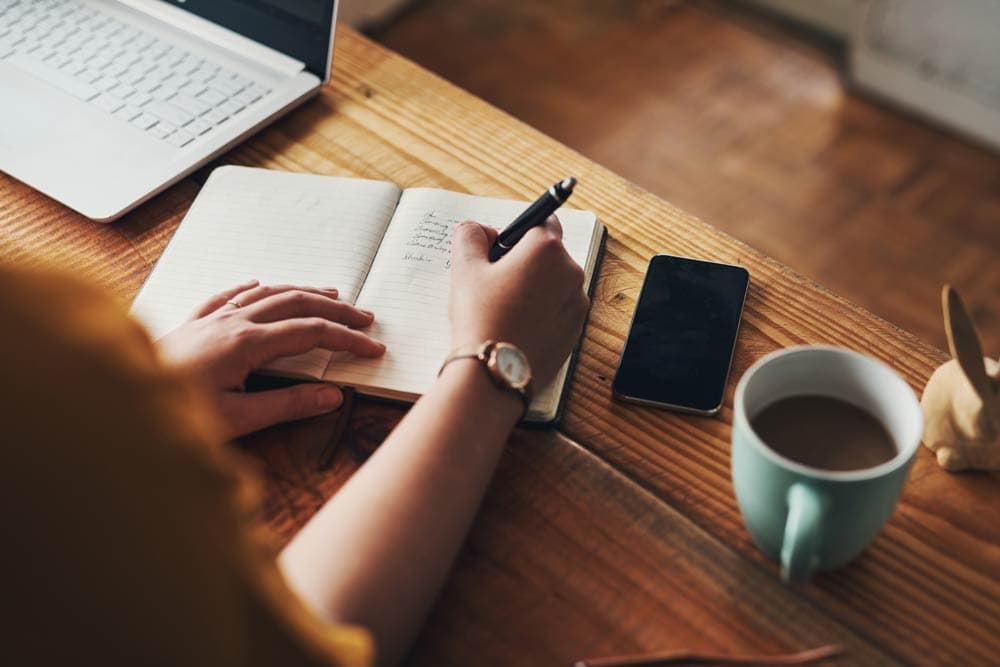 Close-up of a person writing in a notebook beside a laptop and coffee, representing habit stacking techniques to build positive behaviours for ADHD management.