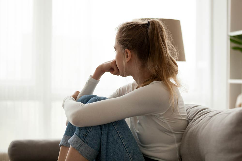 A young woman sitting on a couch looking thoughtful and distressed, symbolizing the emotional pain of rejection sensitivity often experienced with ADHD.