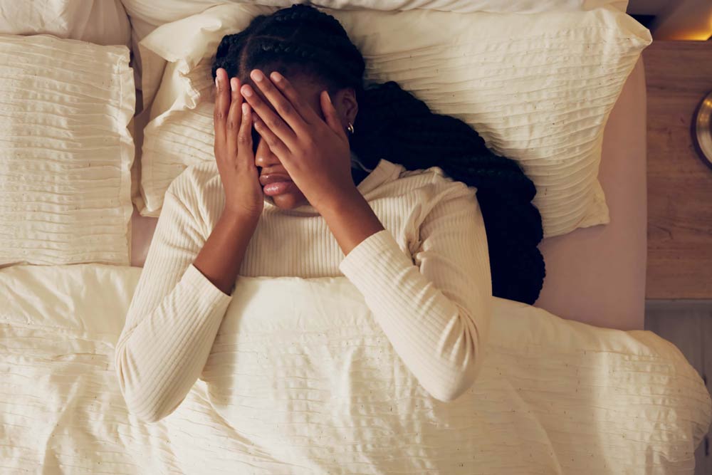 A woman lying in bed covering her face with her hands, representing frustration and exhaustion from ADHD-related sleep difficulties.