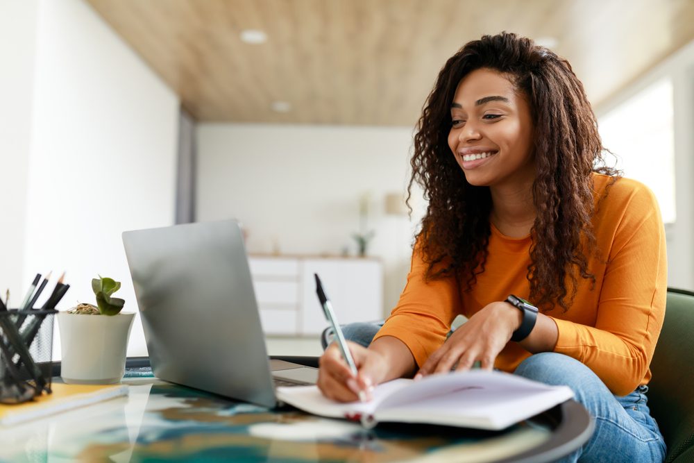 Person at a desk writing dated notes in a small notebook beside a laptop