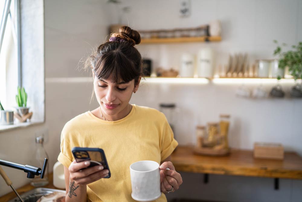 Young woman in a kitchen holding a phone and coffee mug, symbolizing how ADHD symptoms in adults can appear subtle and often go overlooked.