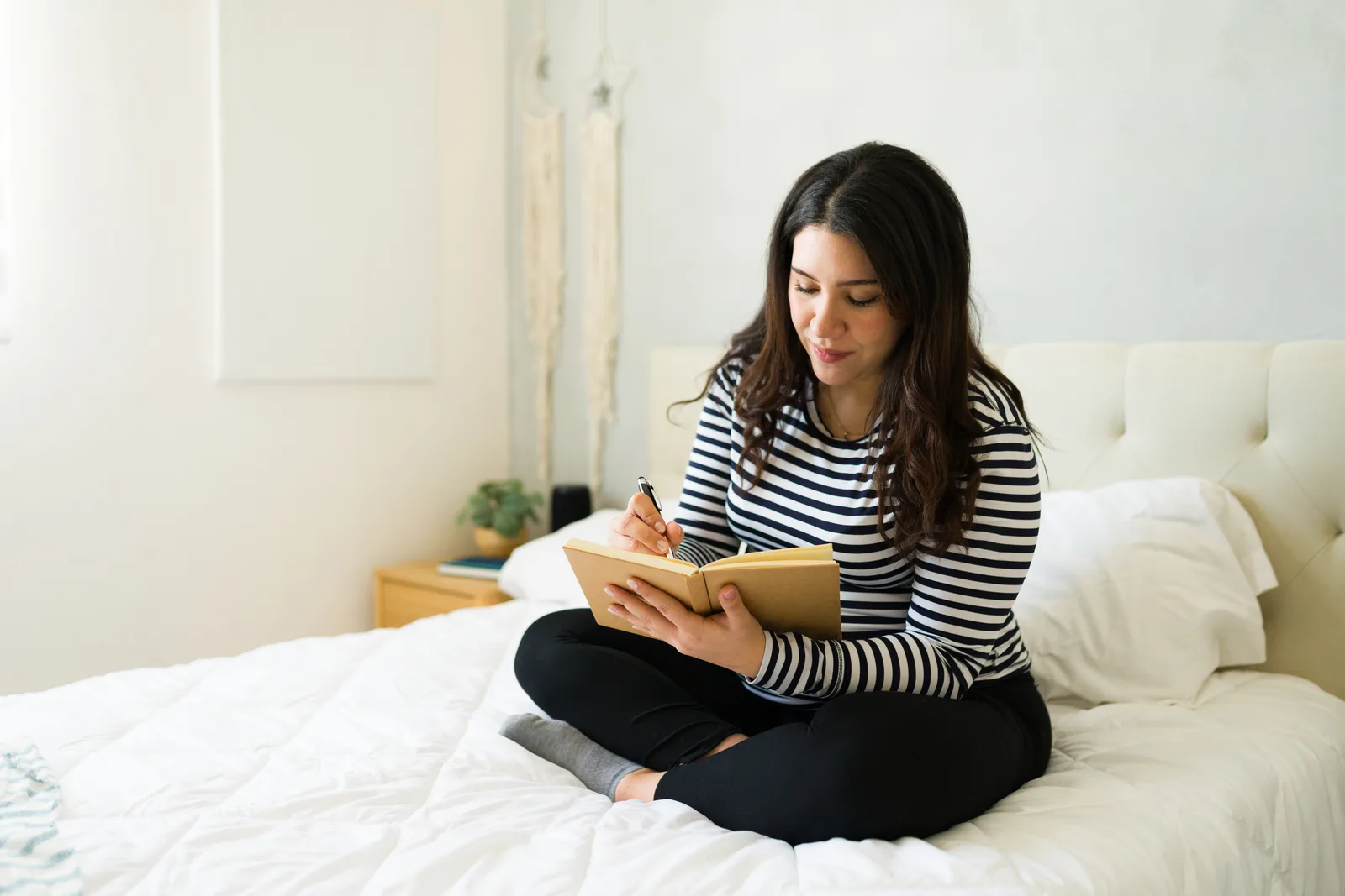 Expectant mother writing a list of questions in a notebook at home in soft morning light.