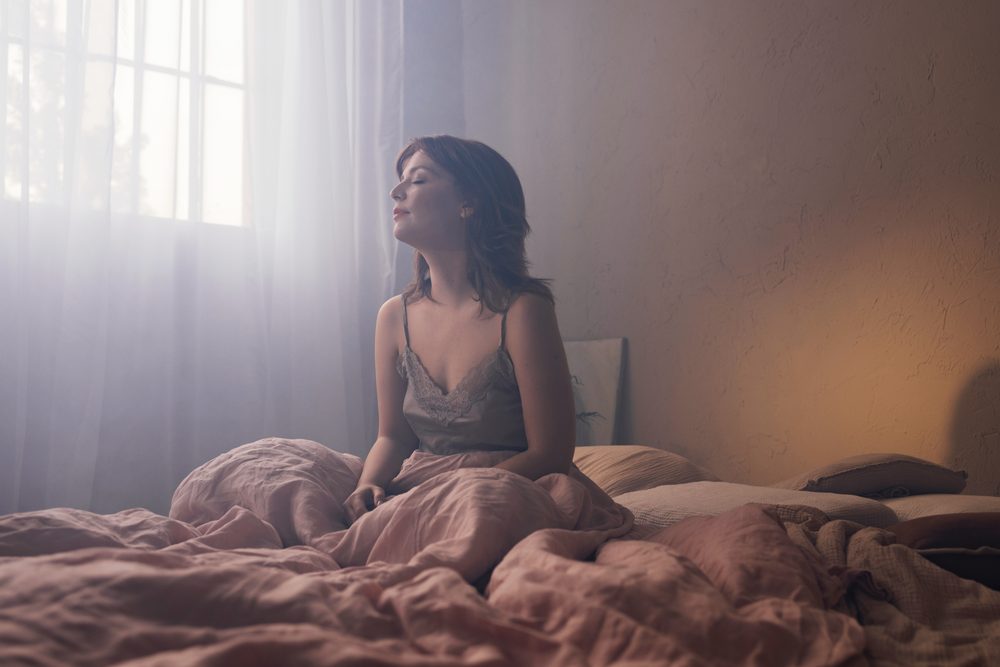 Sunlight through open blinds onto an unmade bed with a glass of water on the nightstand