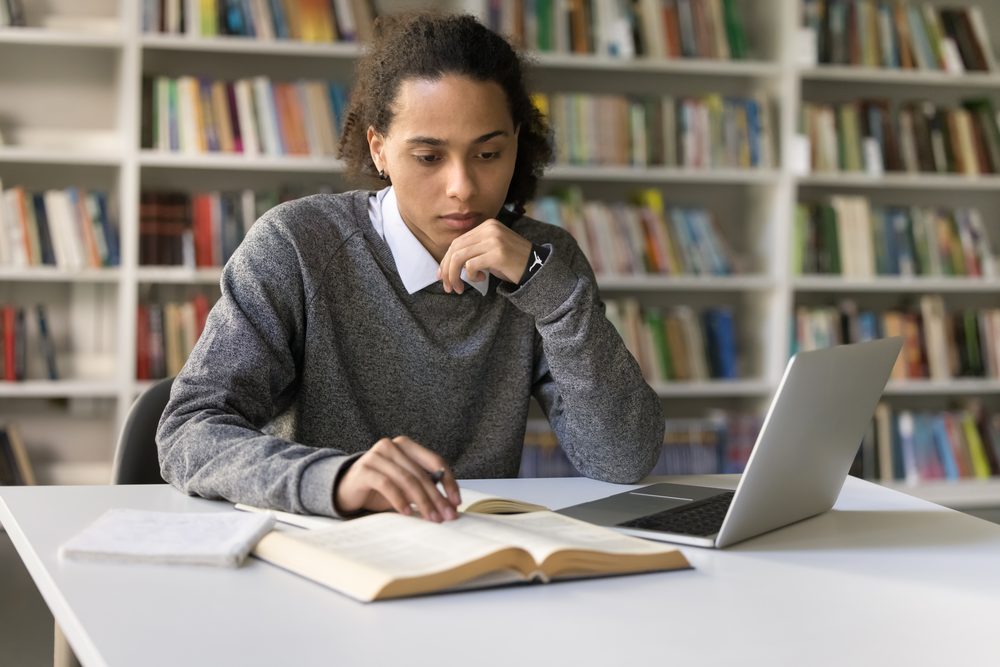 University student at a library table with a laptop, phone and printed syllabus
