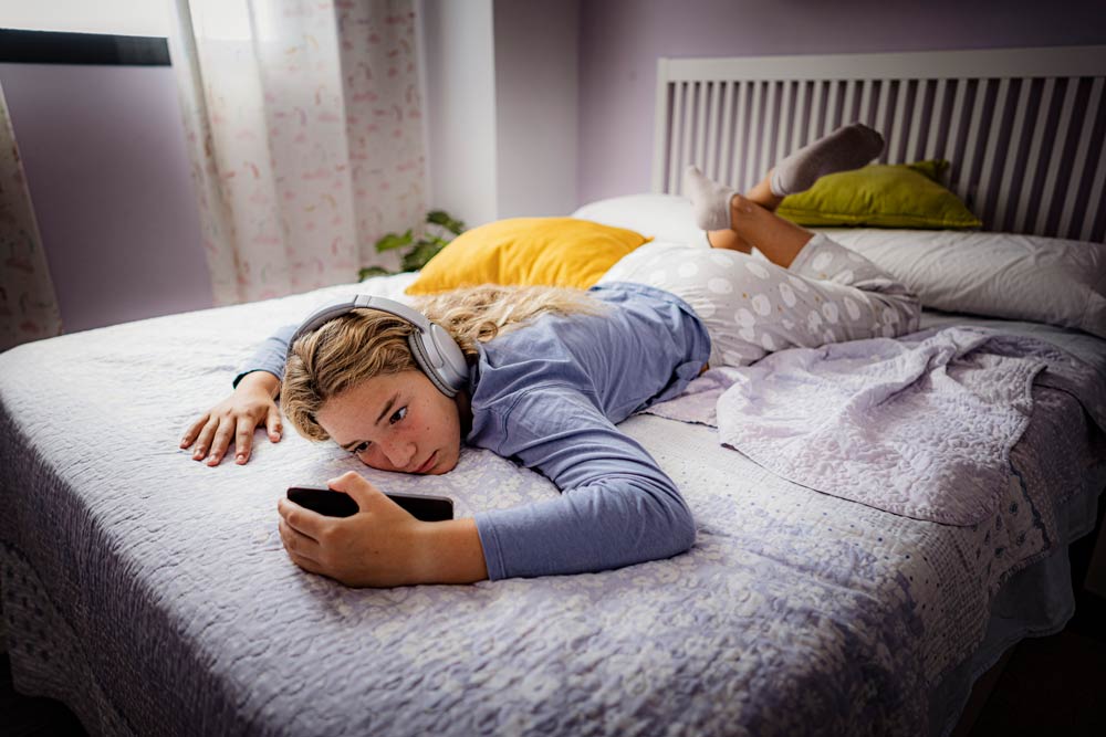 A teenage girl lies on her bed with headphones and a phone, illustrating the challenges teens with ADHD face with executive functioning, focus, and task management.