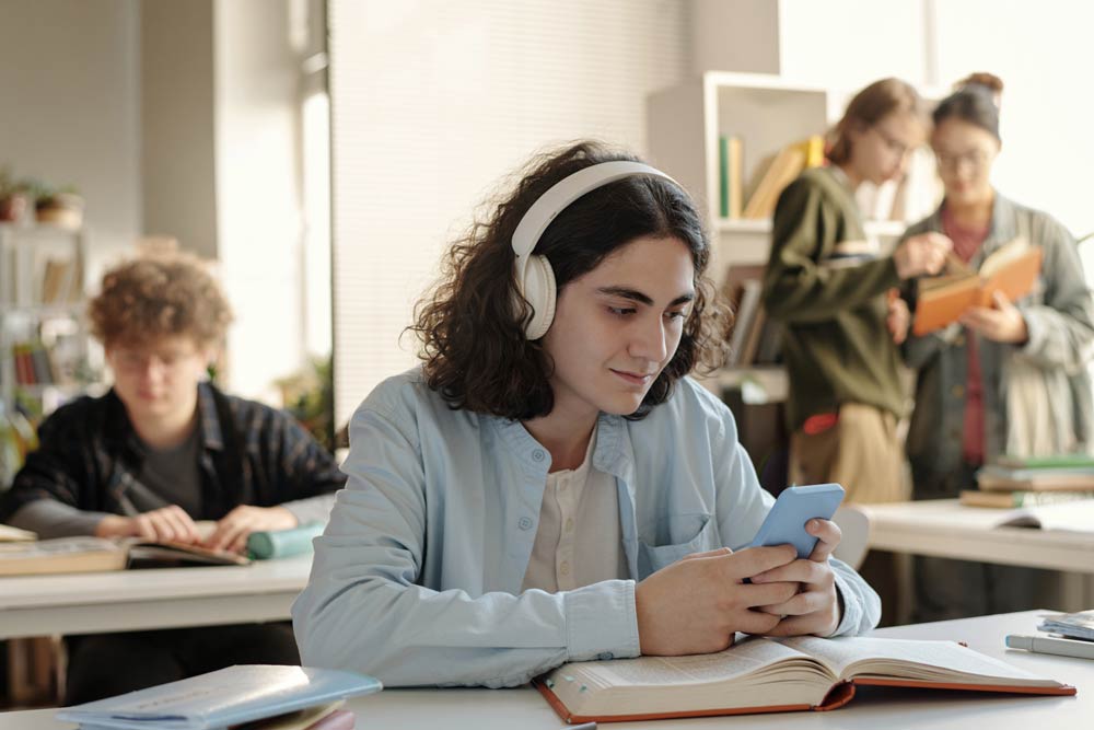 A teenage student wearing headphones looks at a phone during class, symbolizing the challenge of managing distractions for teens with ADHD in school.