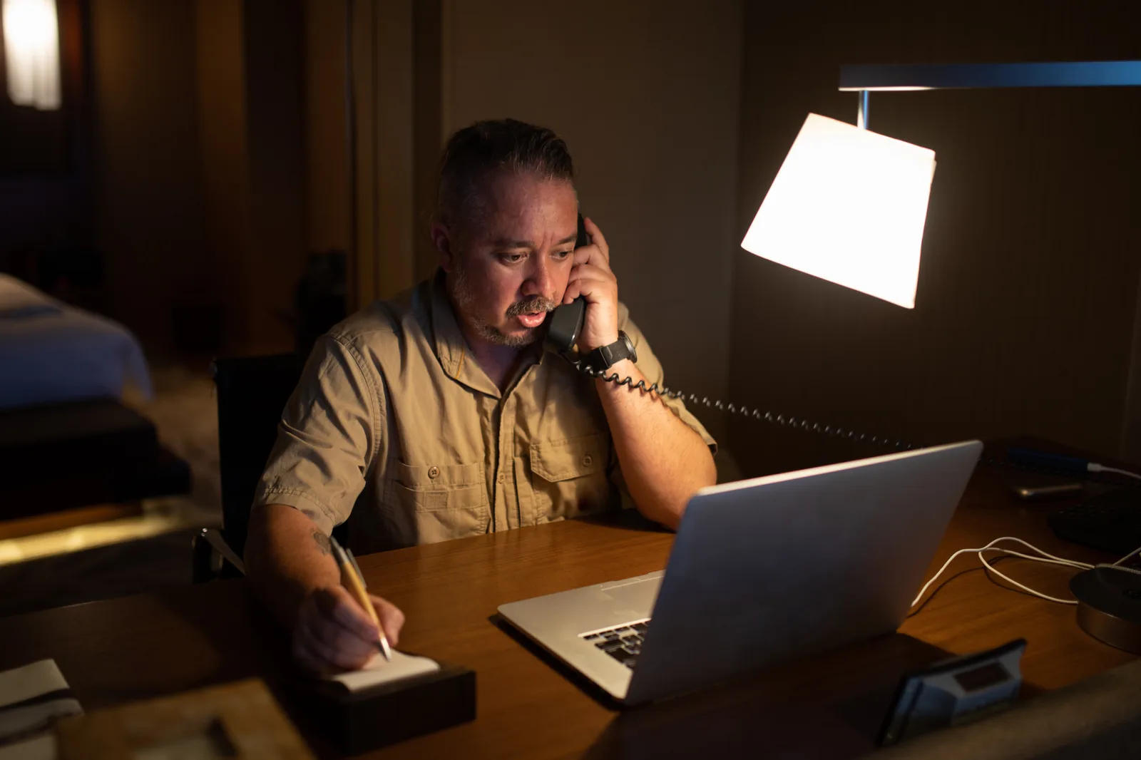 A man on a phone call with his insurance provider, taking notes about his health benefits at a home office desk.