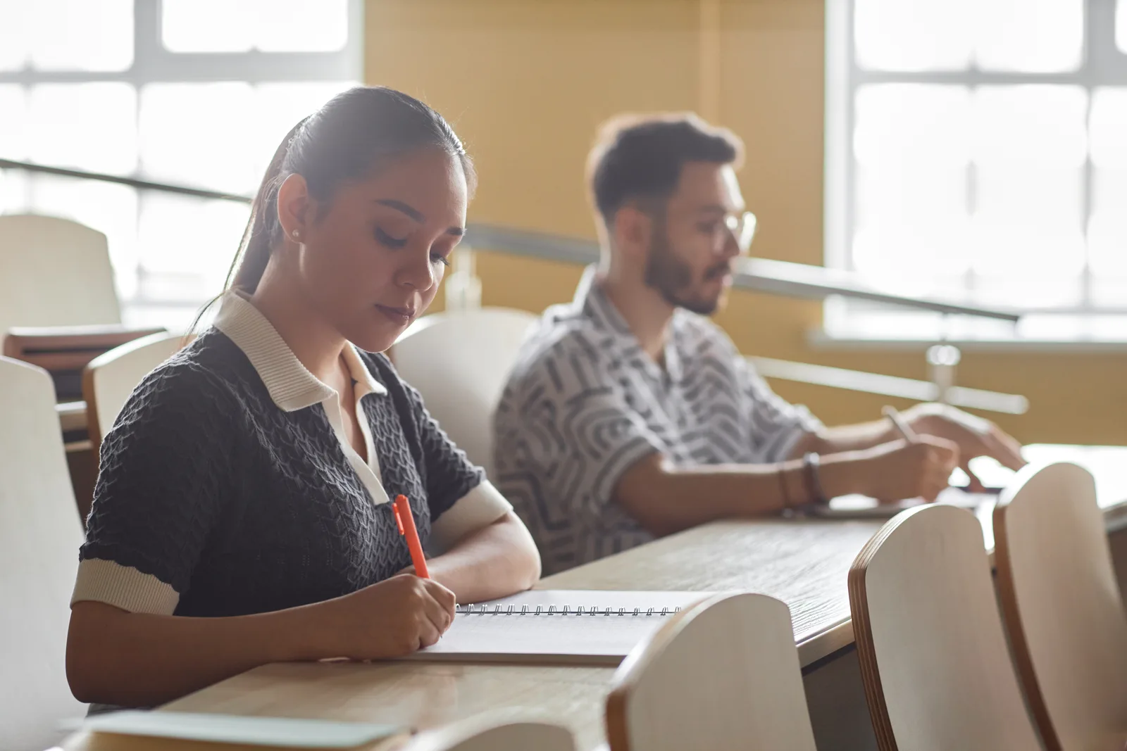 A focused college student writing an exam at a desk by a window in a quiet, distraction-reduced room.