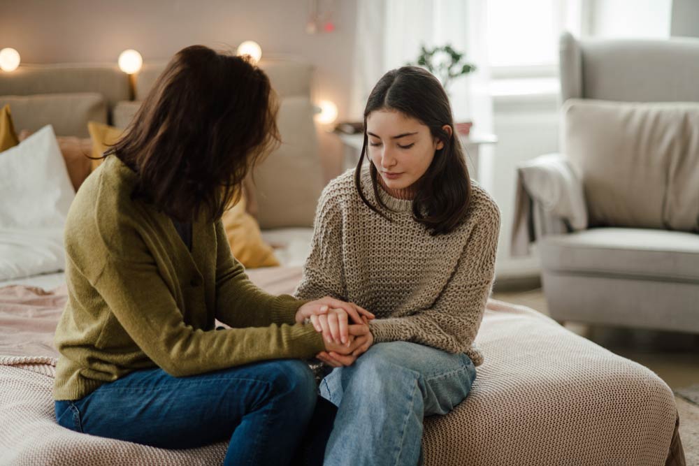 A parent holds their teenager’s hands while talking on the bed, representing open communication and emotional support when explaining ADHD to family members.