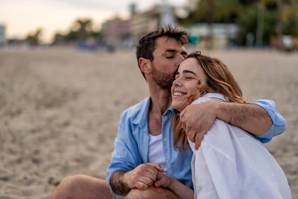 A man kissing a smiling woman on the forehead while holding hands on the beach, symbolizing the effects of ADHD on dating and romantic attraction.