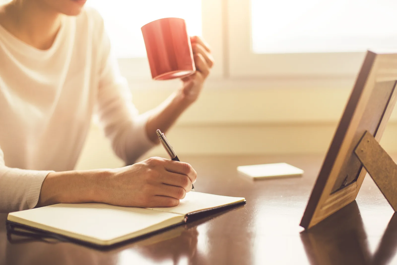 A woman writes preparation notes in a notebook at her desk beside a mug of coffee, getting organized before her ADHD assessment appointment.