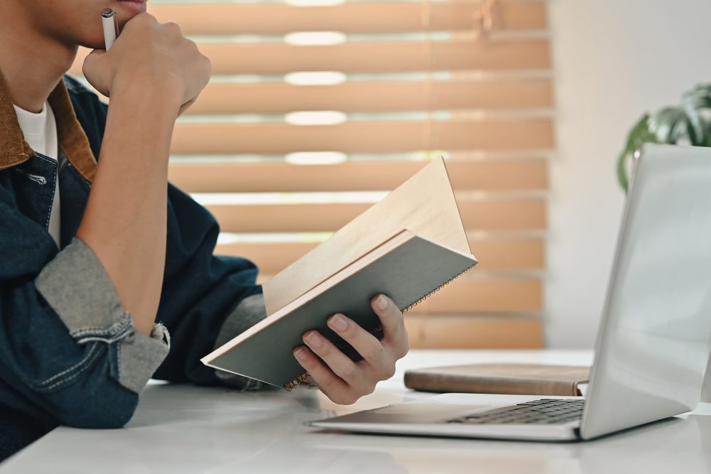 Person at a kitchen table with a laptop, a notebook and a box of desk items