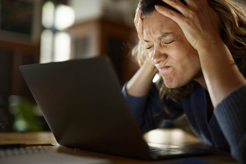 A frustrated woman holding her head in front of a laptop, representing the challenges of anger and emotional dysregulation experienced by individuals with ADHD.