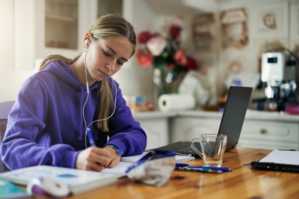 A teenage student in a purple hoodie studies at home with earphones in, writing notes beside a laptop, representing ADHD homework strategies and focus support.