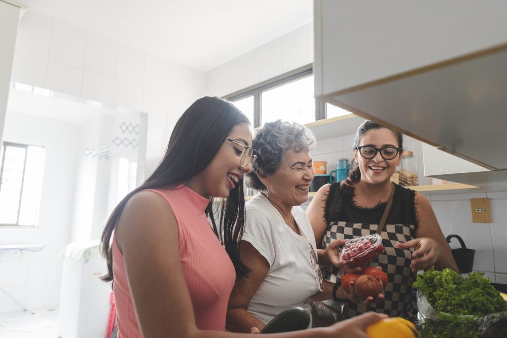Mother at a cluttered kitchen counter holding a phone while two children eat breakfast