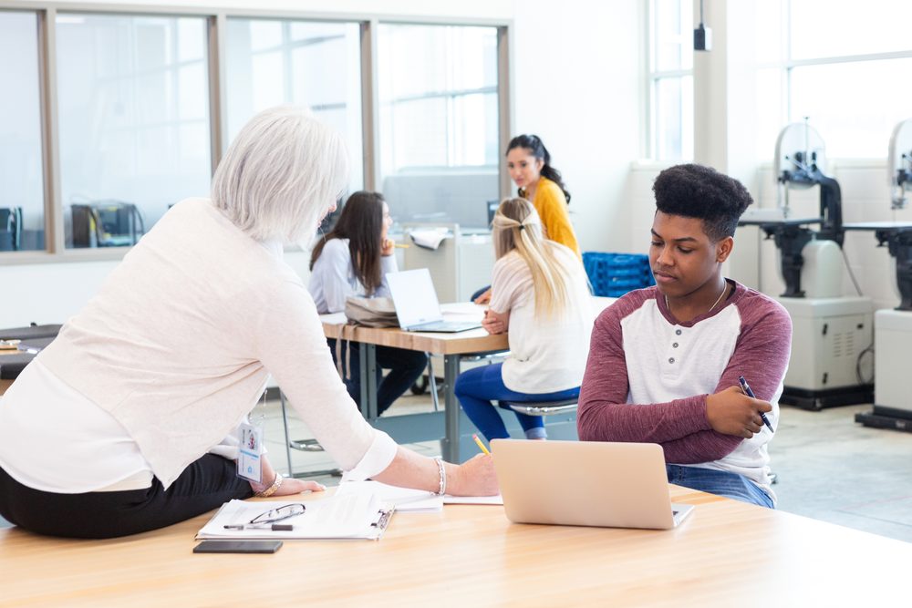 Student seated at a cleared desk facing a laptop webcam with student ID beside the keyboard