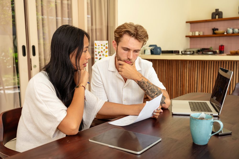 Man at a kitchen table looking at a laptop screen while his partner stands behind him