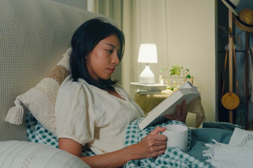 Woman reading a book in bed with a warm drink, representing healthy bedtime routines and the importance of sleep hygiene for managing ADHD symptoms.