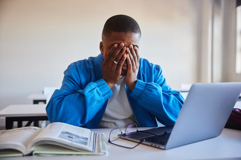 A stressed college student with ADHD covering their face while studying with a laptop and textbooks, representing the emotional and academic toll of burnout.