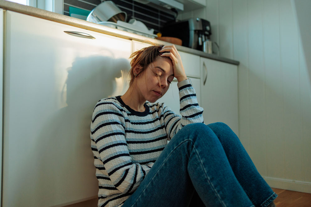 A woman sitting on the floor with her head in her hand, looking exhausted, representing the emotional and physical signs of ADHD-related burnout.