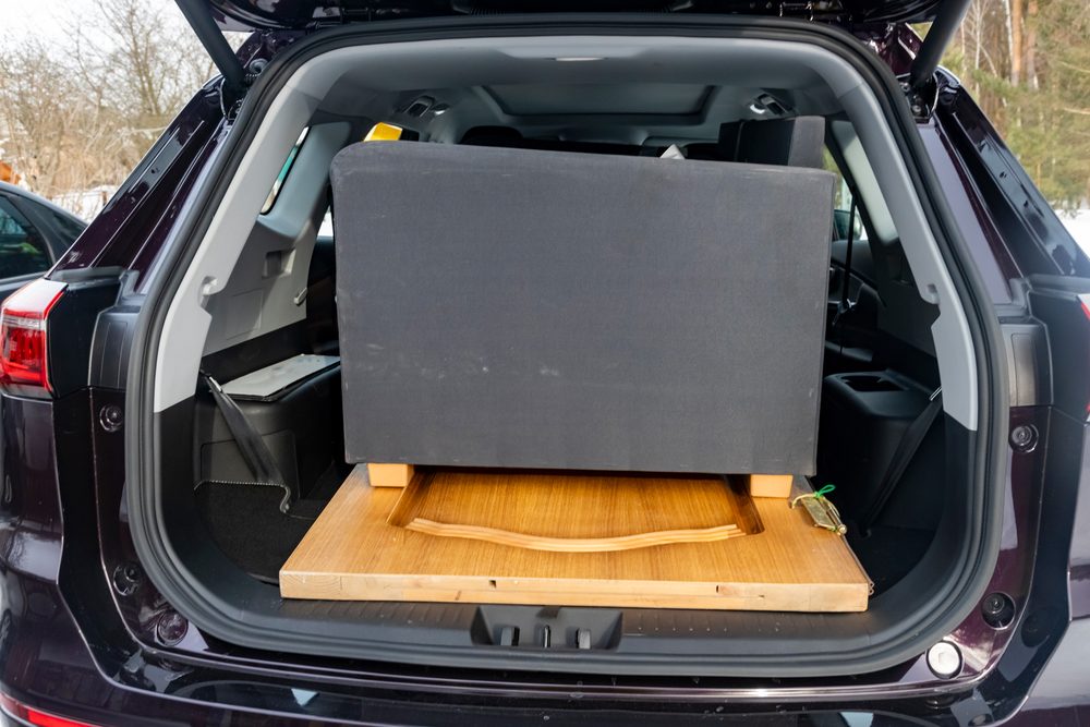 Driver loading a cardboard return parcel into a car trunk outside a strip mall