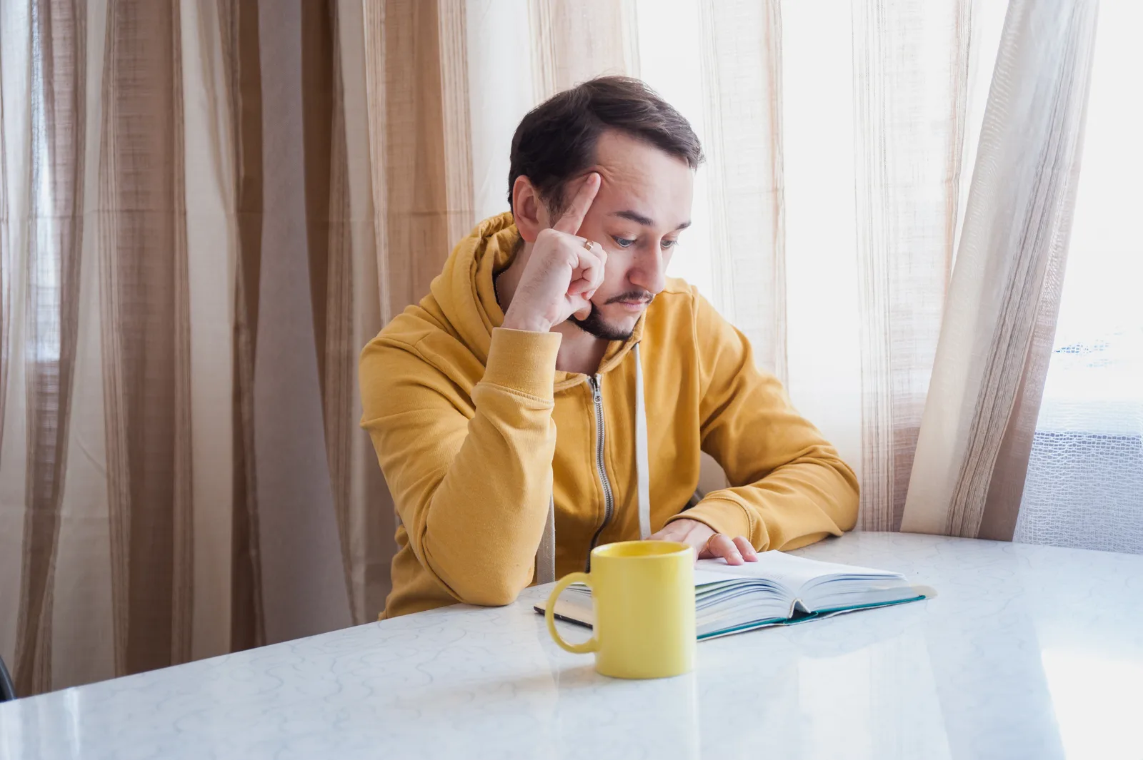 Thoughtful man reading and taking notes in a notebook at a sunny desk with a mug beside him.