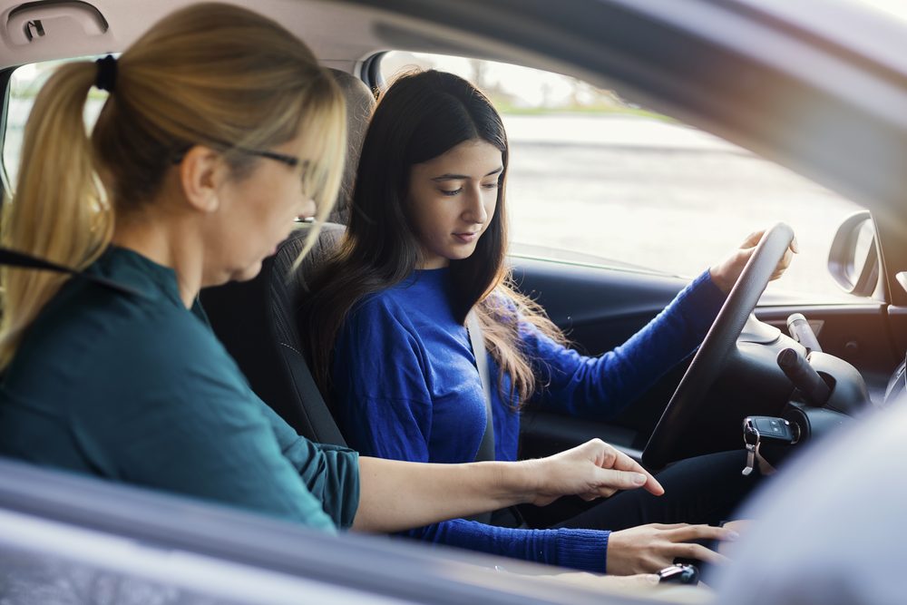 Teenage girl at the wheel of a parked car, parent in the passenger seat pointing ahead