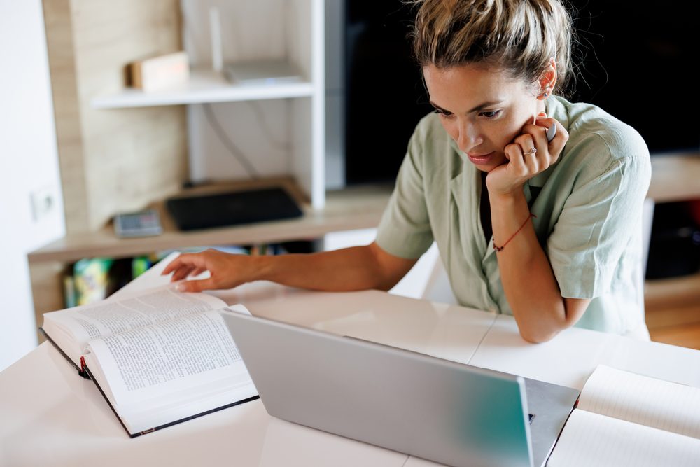 Graduate student at a desk piled with journal articles, typing on a laptop by a window