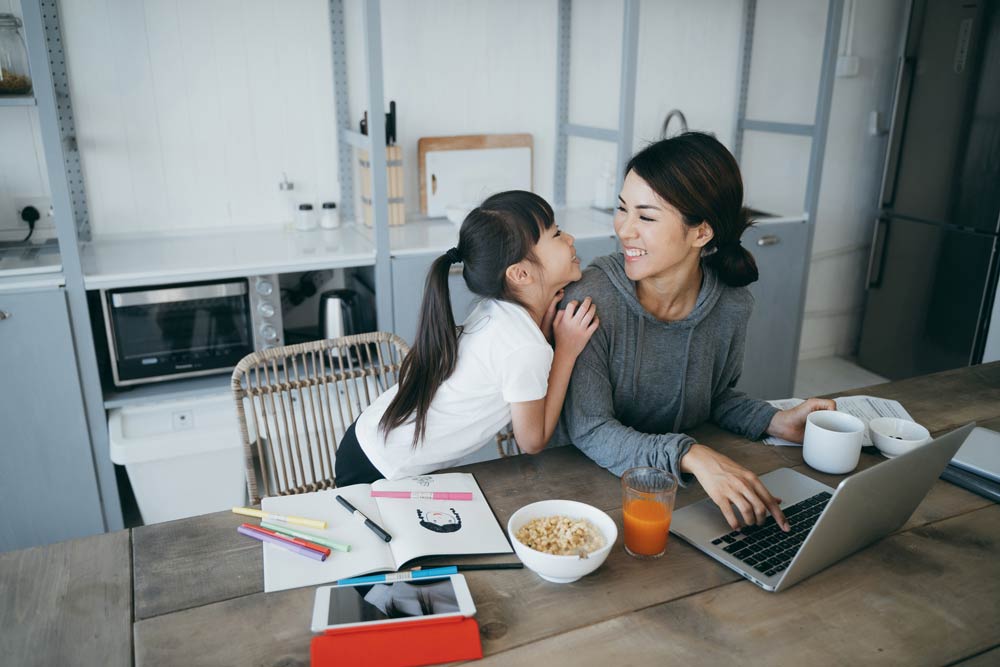 Mother with ADHD smiling at her young daughter while working from home on a laptop, representing balance between parenting and managing ADHD.