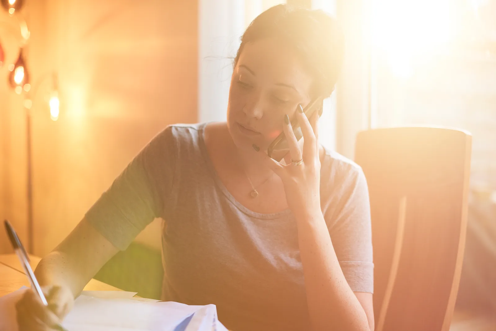 Woman on the phone reviewing paperwork at a kitchen table with a coffee mug in warm afternoon light.