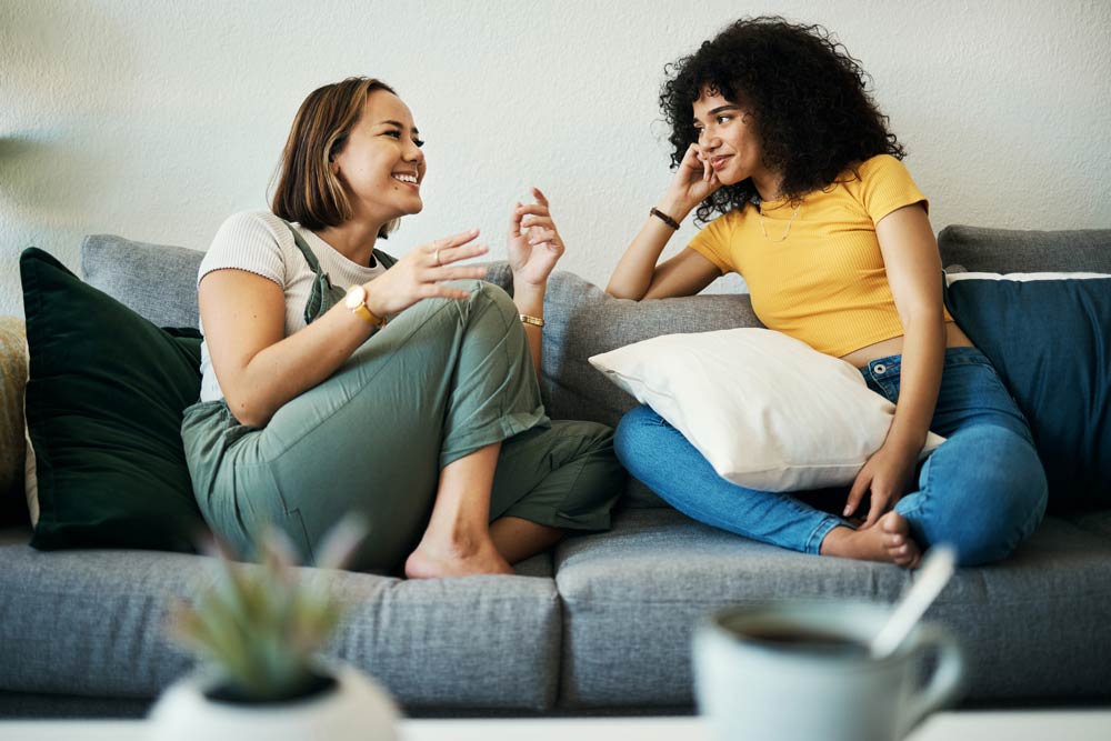 Two women sit on a couch talking and smiling, representing how ADHD affects communication in women and the importance of strategies for healthier relationships.