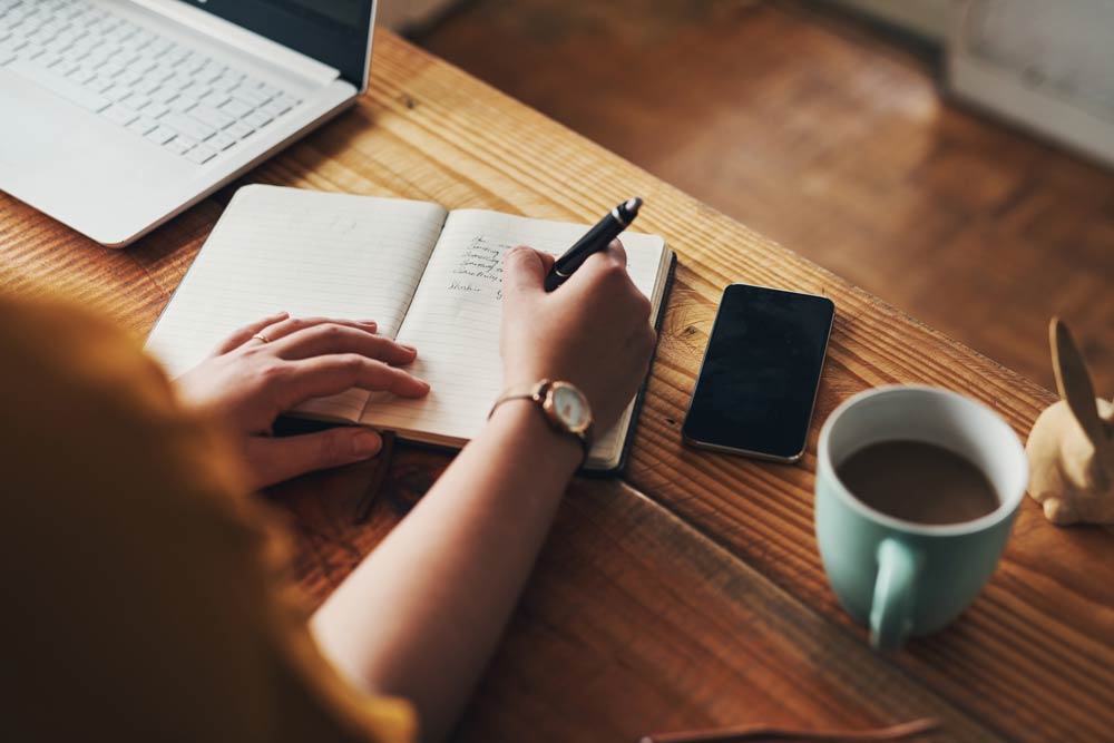 Close-up of a person writing in a notebook beside a laptop and coffee, representing habit stacking techniques to build positive behaviours for ADHD management.
