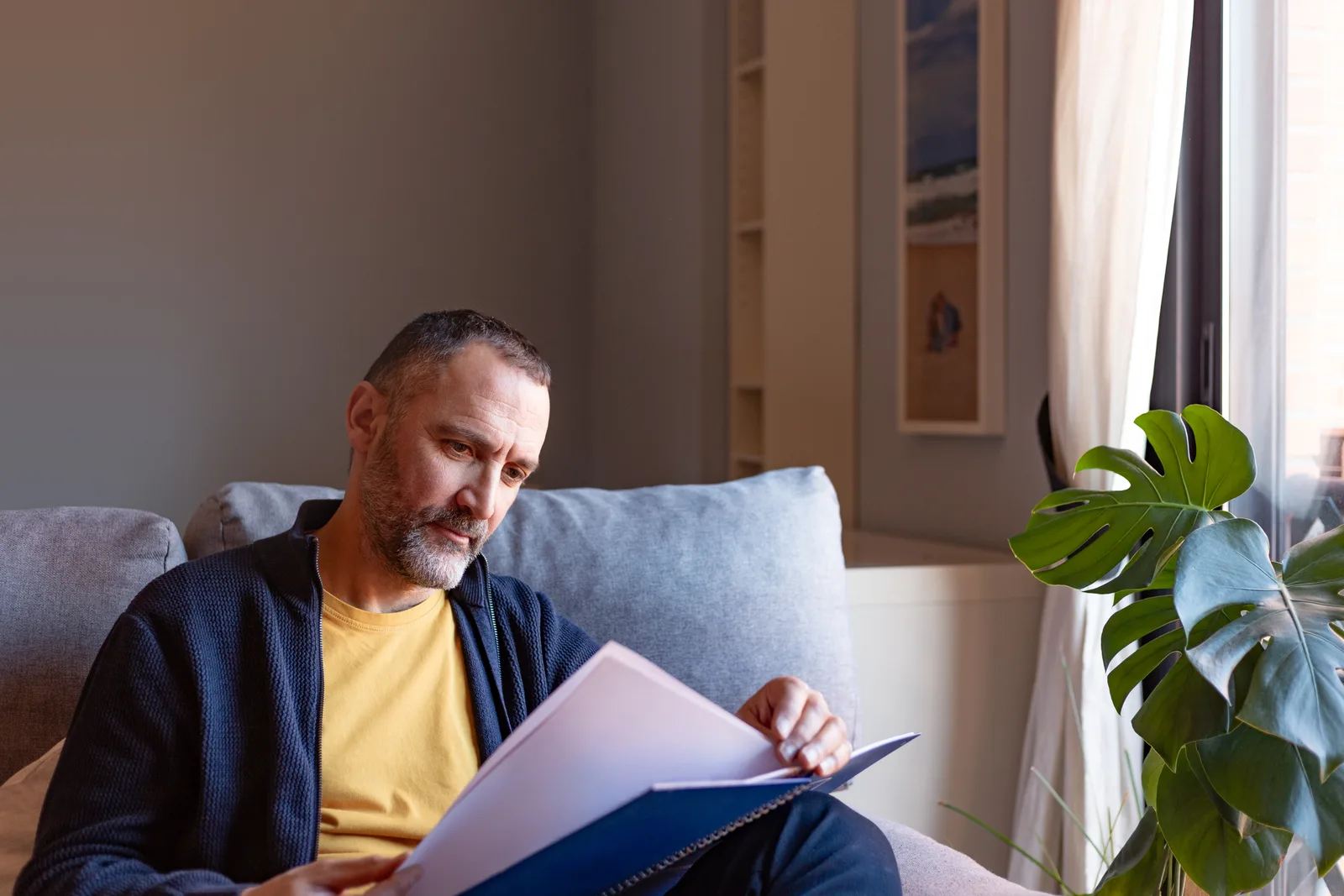 Man at home on a sofa organizing personal notes and school records into a folder, preparing ahead of an upcoming ADHD assessment.