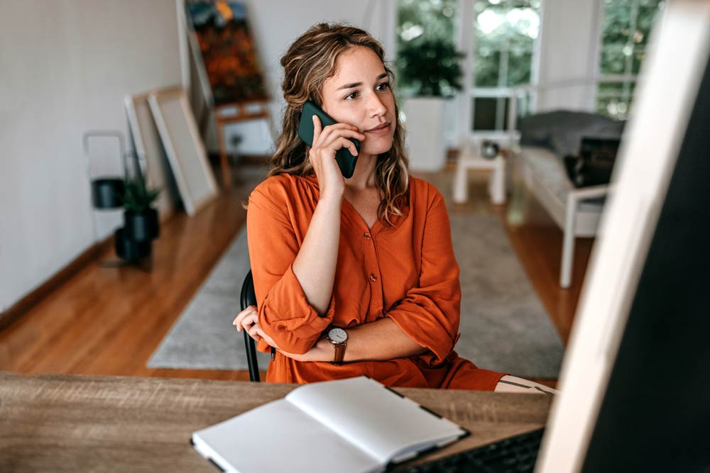Young woman in an orange shirt talking on the phone while working from home, representing the overlooked and underdiagnosed signs of ADHD in women.