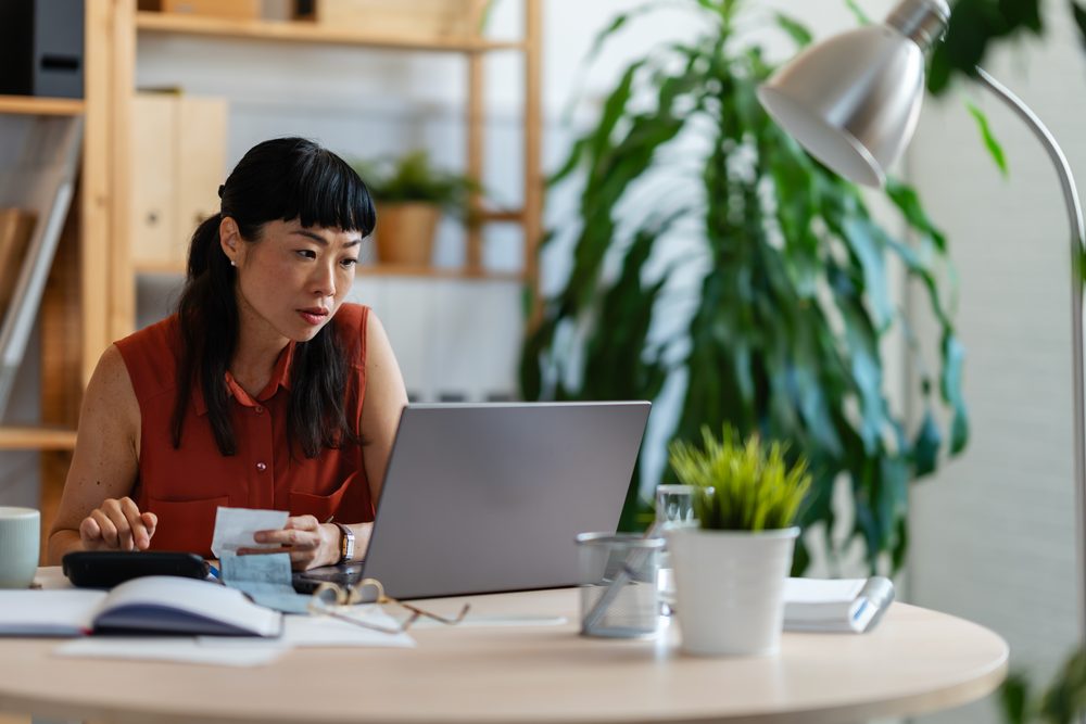 Person at a small home desk by a window with a laptop, notebook and coffee mug